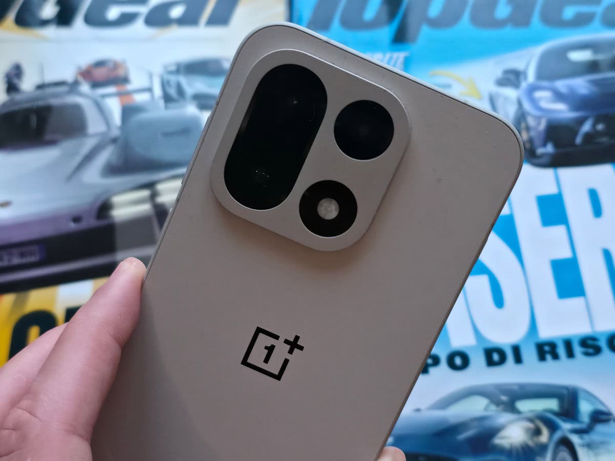 Alt: Primo piano di uno smartphone OnePlus 15 con tripla fotocamera posteriore, sfondo di auto da corsa e poster di automobilismo, in evidenza tecnologia e dispositivi mobili.