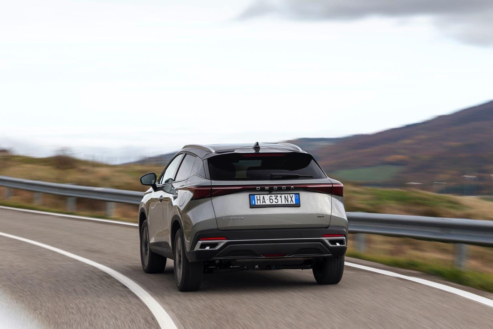 Auto SUV Omoda 5 SHS-S in movimento su strada di montagna con sfondo paesaggio naturale, immagine dinamica di veicolo elettrico, design futuristico, tecnologia avanzata, auto compatta di lusso, notizie automotive.