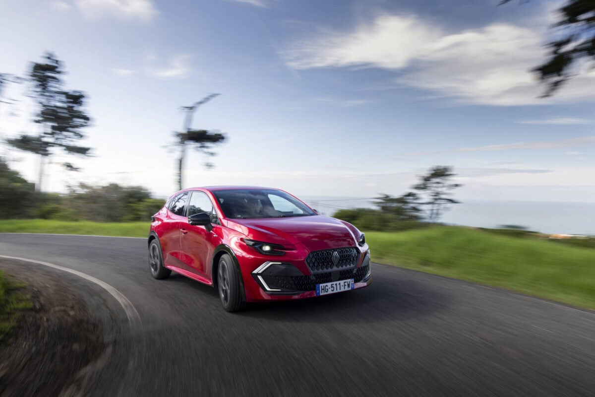 Auto Nuova Renault Clio 2026 rossa in azione su curva di strada panoramica, sfondo naturale con cielo nuvoloso, perfetta per immagini di veicoli sportivi e test drive.