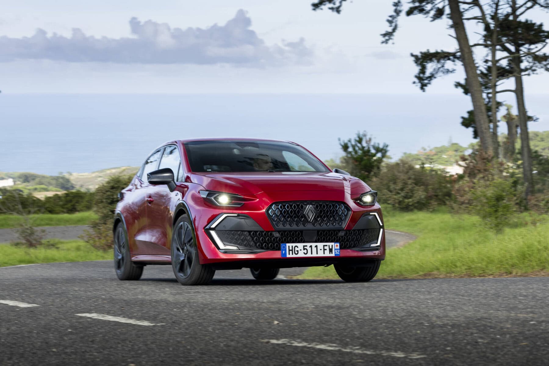 Auto sportiva Nuova Renault Clio 2026 in colore rosso acceso che percorre una strada con vista mare e natura, in un ambiente di viaggio e avventura.