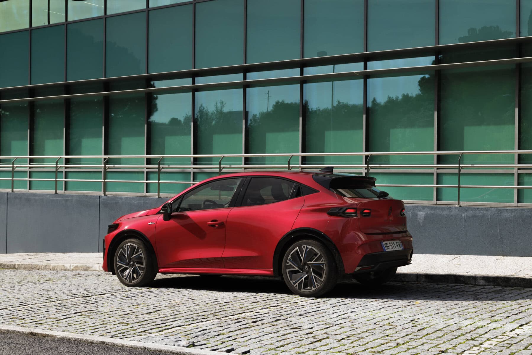 Auto Nuova Renault Clio 2026 rossa moderna in città, nello stile delle auto elettriche di nuova generazione, parcheggiata su strada con edificio in vetro sullo sfondo.