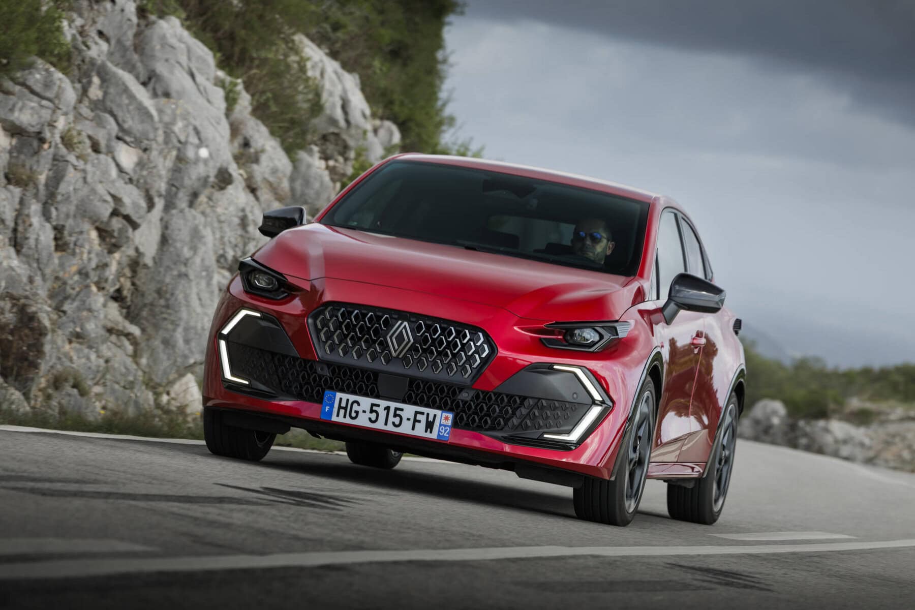 Auto Nuova Renault Clio 2026 rossa in movimento su strada di montagna con sfondo naturale e cielo nuvoloso.