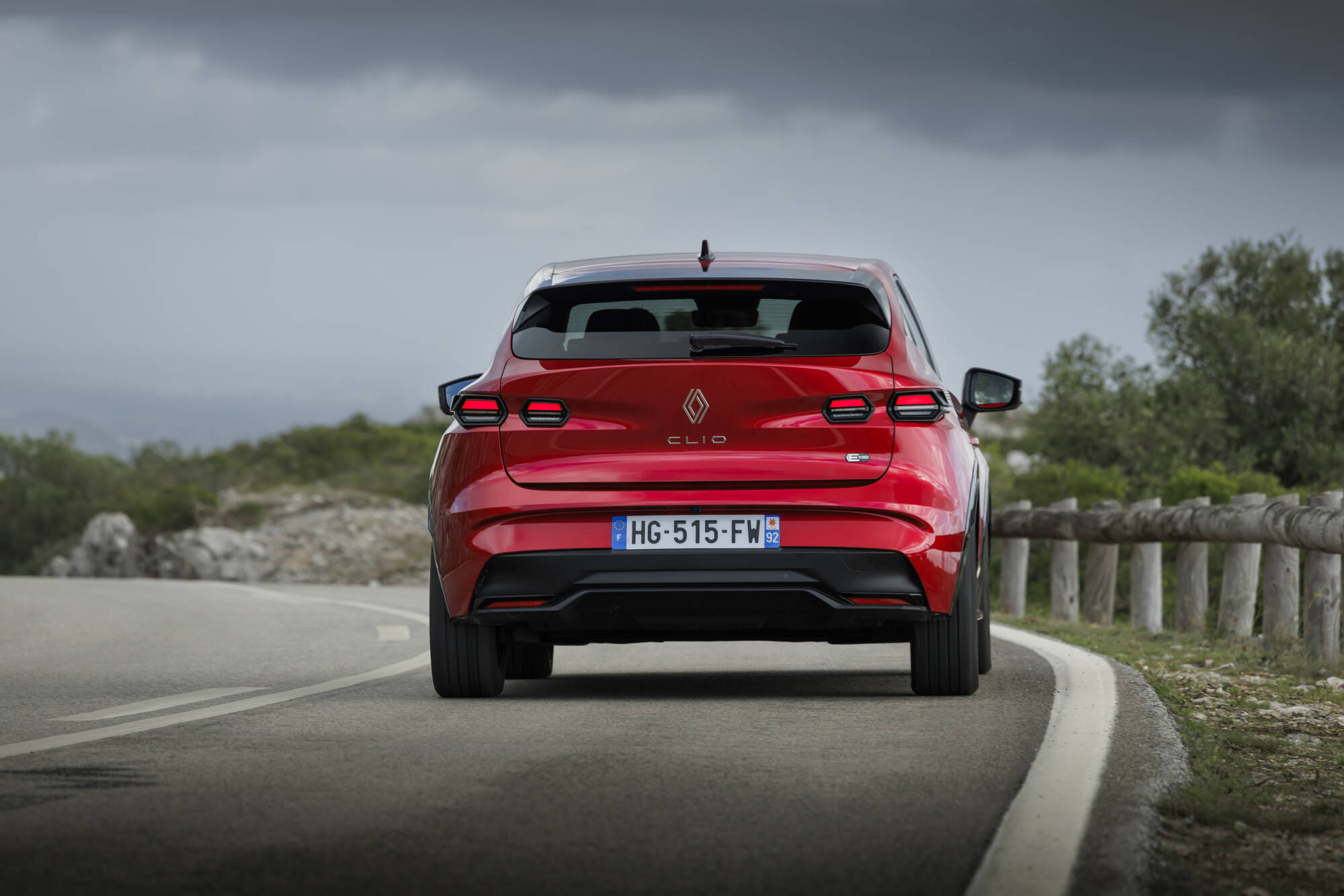 Auto Nuova Renault Clio 2026 rossa vista da dietro su strada con paesaggio naturale, design moderno e sostenibilità, modello Renault Clio E-Tech Hybrid, esempio di auto ecologica e innovativa.