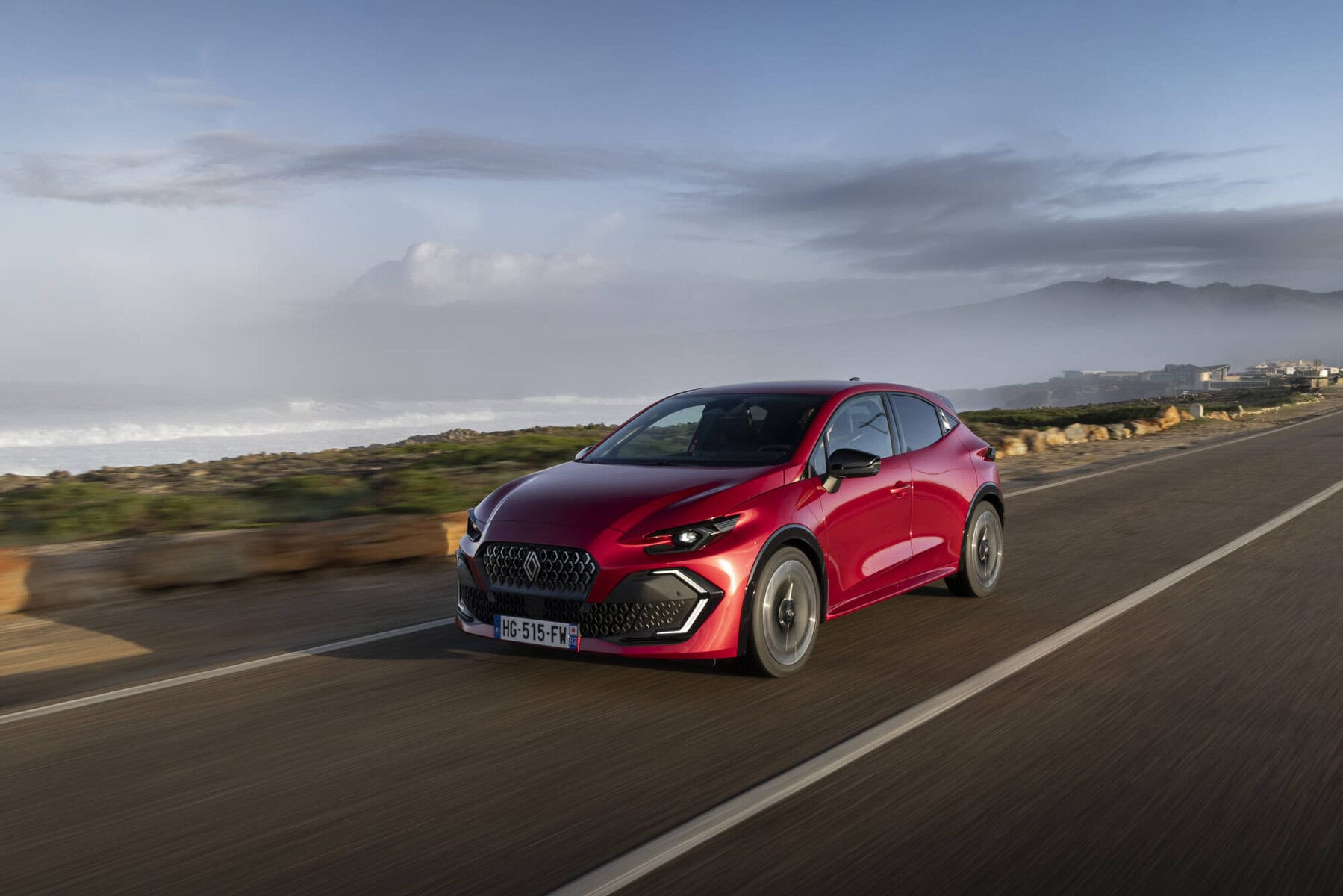 Auto Nuova Renault Clio 2026 moderna rossa in movimento lungo la costa con montagne sullo sfondo, perfekte per appassionati di automobili e viaggi on the road.