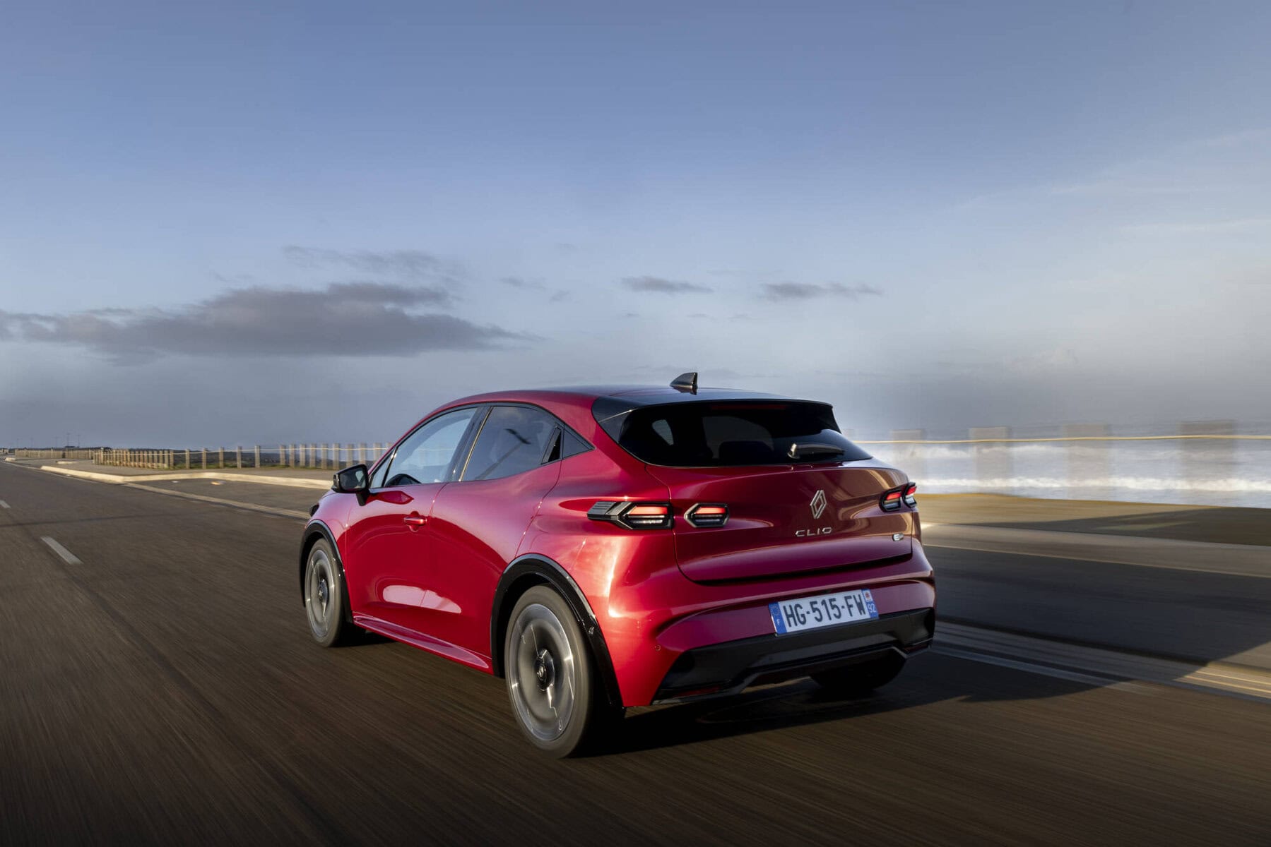 Auto elettrica Nuova Renault Clio 2026 rossa in movimento su strada asfaltata, paesaggio costiero, cielo sereno, veicolo sostenibile, tecnologia avanzata, auto moderna, zero emissioni, design aerodinamico.
