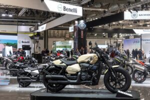 Moto Benelli moderna in esposizione a una fiera motociclistica, con vari modelli di moto attorno e pubblico interessato, stand Benelli ben visibile sullo sfondo.