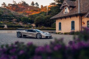 Auto Gordon Murray S1 LM superleggera moderna parcheggiata vicino a una casa in stile cottage, con paesaggio collinare e giardino fiorito al tramonto, immagini di lusso e alta performance.