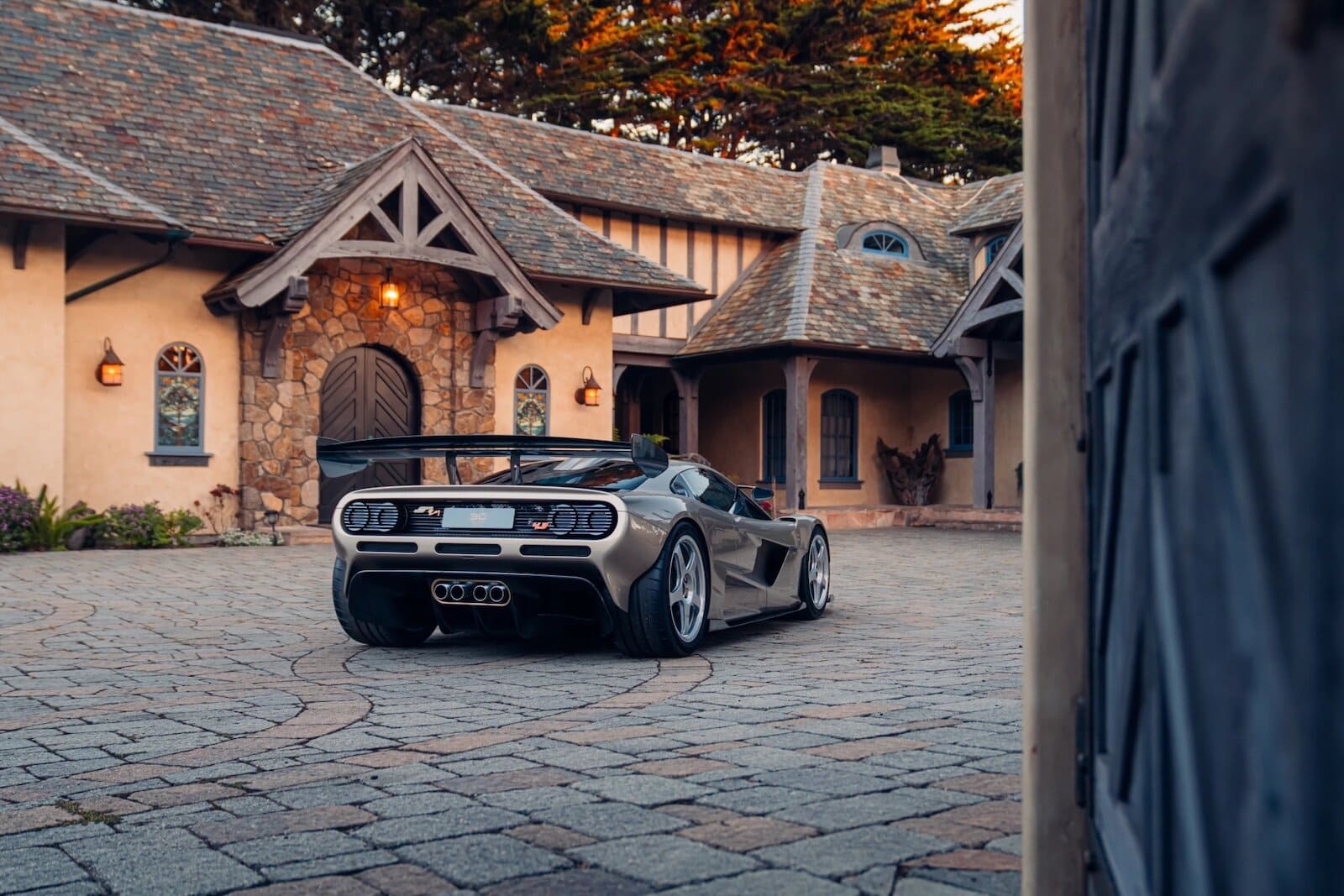 Auto sportiva Gordon Murray S1 LM parcheggiata davanti a una casa di campagna in stile rustico con pietra e legno, su strada di pietre.