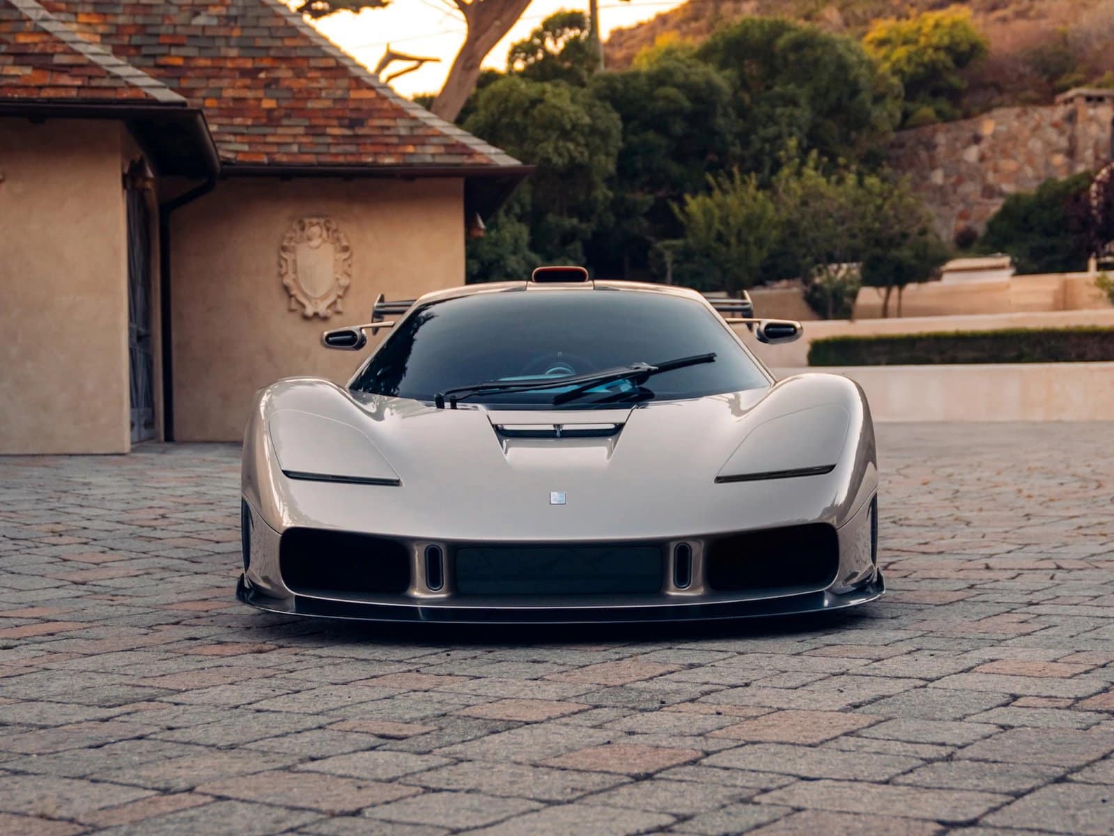 Auto sportiva Gordon Murray S1 LM color argento, in primo piano su strada di pietra, vista frontale, con paesaggio di case e colline sullo sfondo.