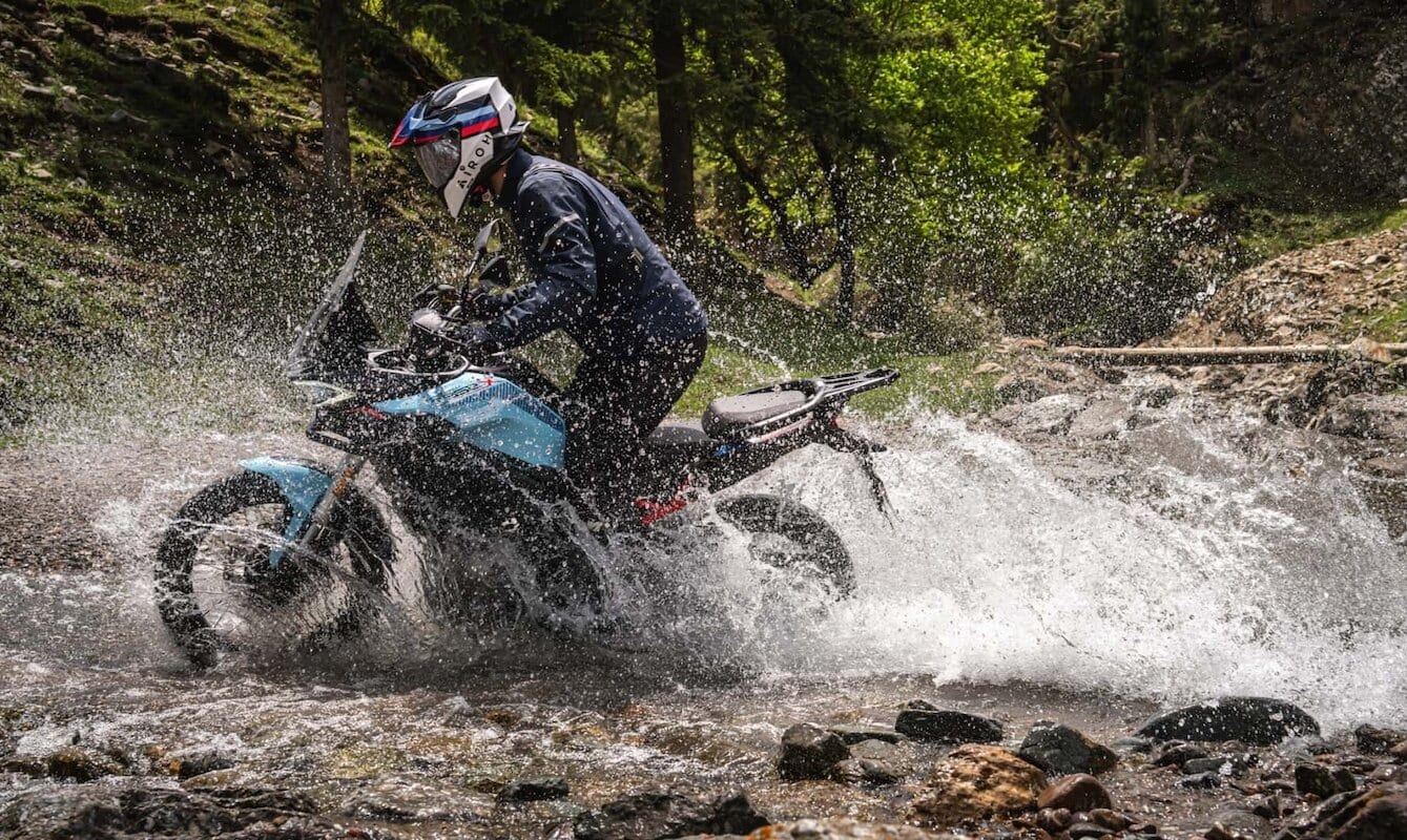 Motociclista con casco e abbigliamento sportivo su moto Morbidelli T352X attraversa un torrente in un'area naturale ricca di alberi.