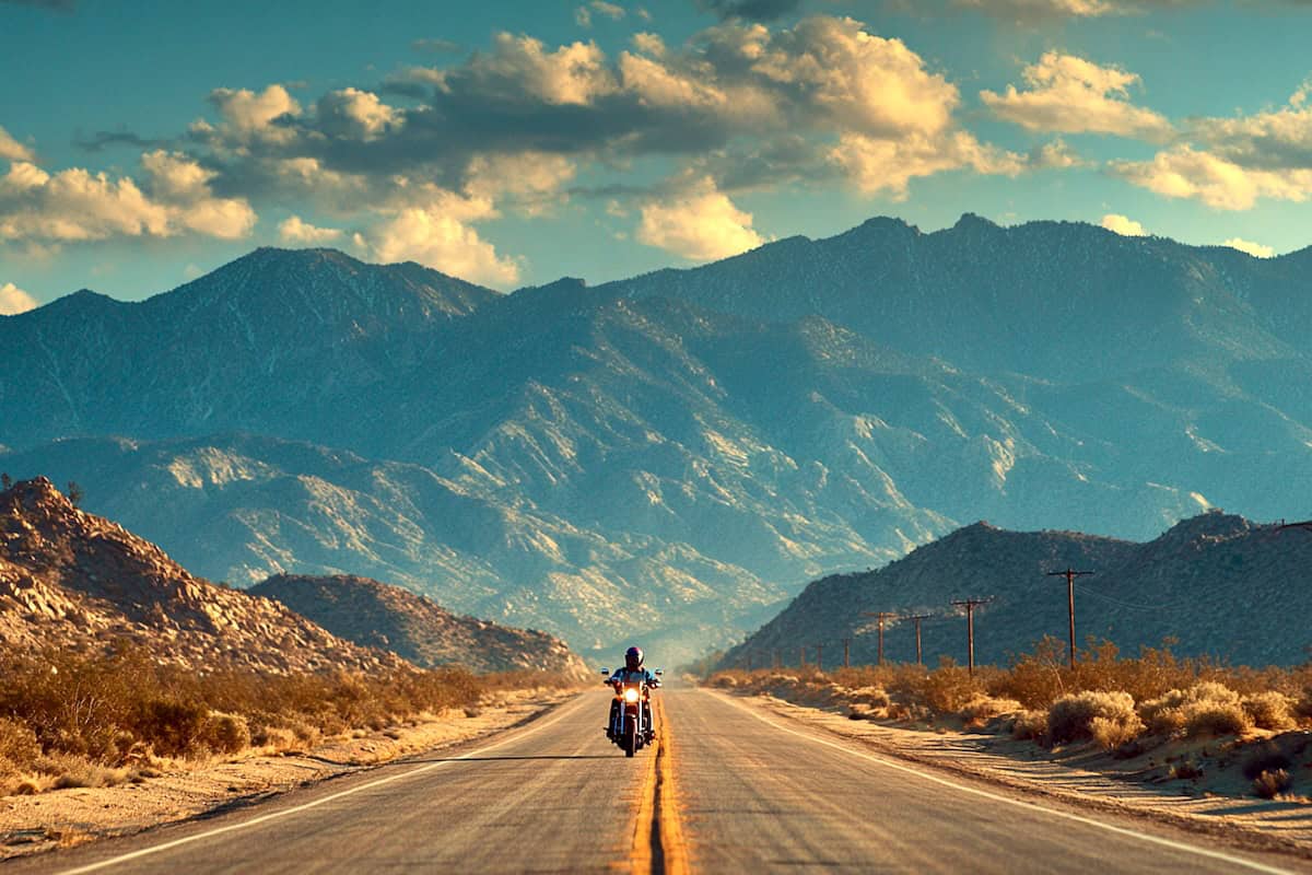 Un motociclista attraversa una strada deserta nel paesaggio montano del deserto, godendosi un viaggio panoramico in un ambiente selvaggio e libero, perfetto per gli appassionati di motociclismo.
