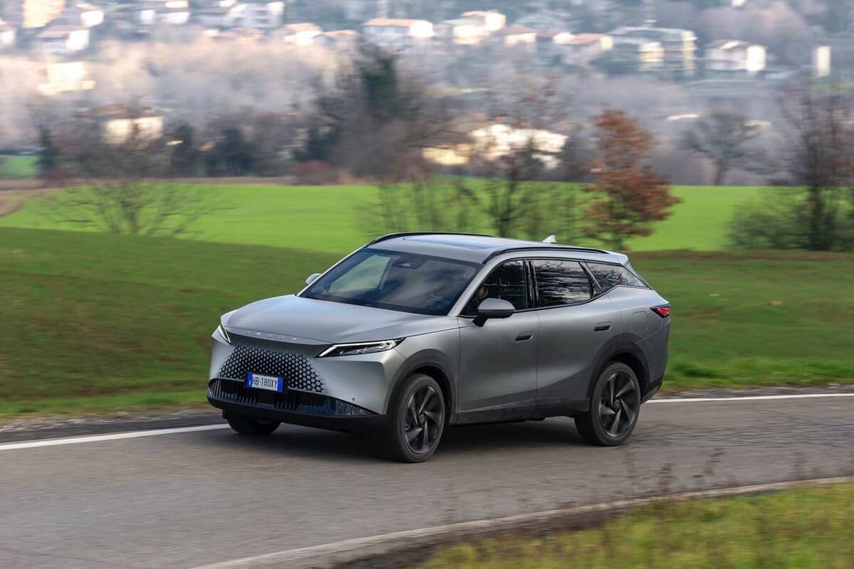 SUV Omoda 7 SHS-P argento, curva su strada di campagna, vista laterale, paesaggio collinare sullo sfondo.