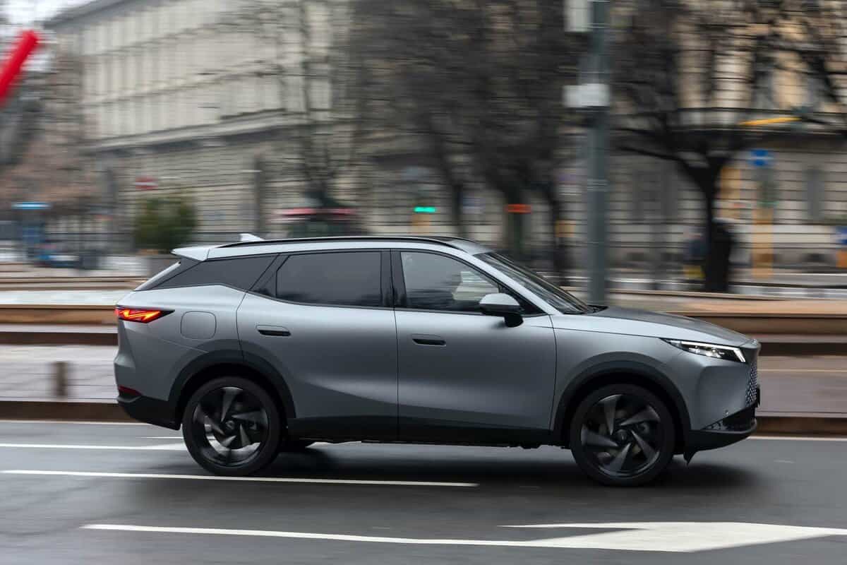 Omoda 7 SHS-P in movimento su strada cittadina, foto laterale, edifici sullo sfondo.