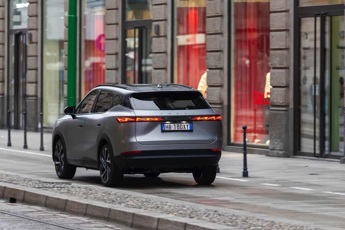 Omoda 7 SHS-P grigio su strada cittadina, visto di tre quarti posteriore, vicino a vetrine negozi.