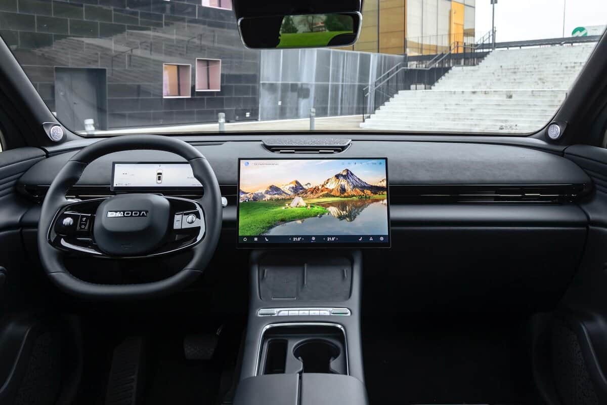 Interni Omoda 7 SHS-P: volante, cruscotto e display centrale ampio, vista dall'abitacolo, edificio in sfondo.