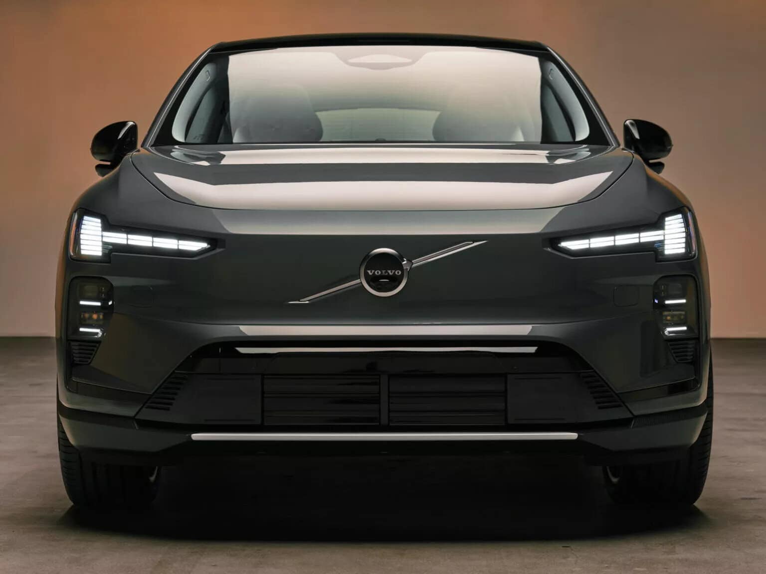 Volvo EX60 grigia, vista frontale, fari a LED accesi, in studio illuminato.