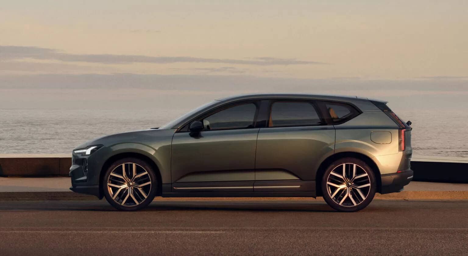 Volvo EX60 grigio al tramonto, vista laterale, vicino al mare su strada costiera.