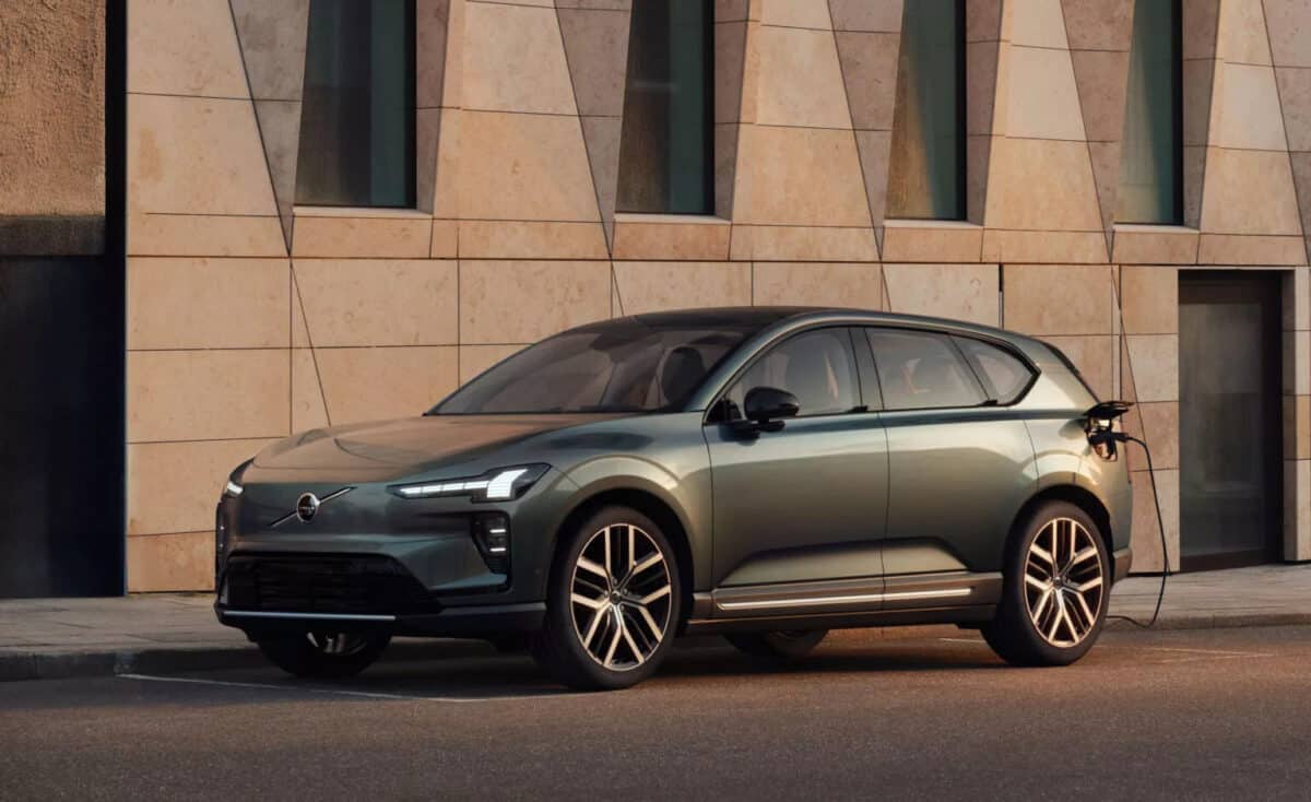 Auto elettrica Volvo EX60 verde in carica, vista laterale su strada urbana vicino a un edificio moderno.