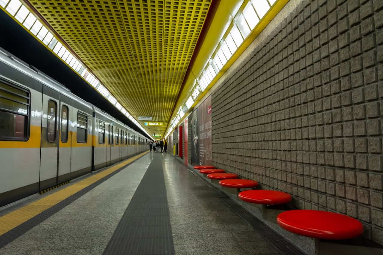 Stazione metropolitana Linea 3 di Milano con sedili rossi e mura di mattoni grigi.