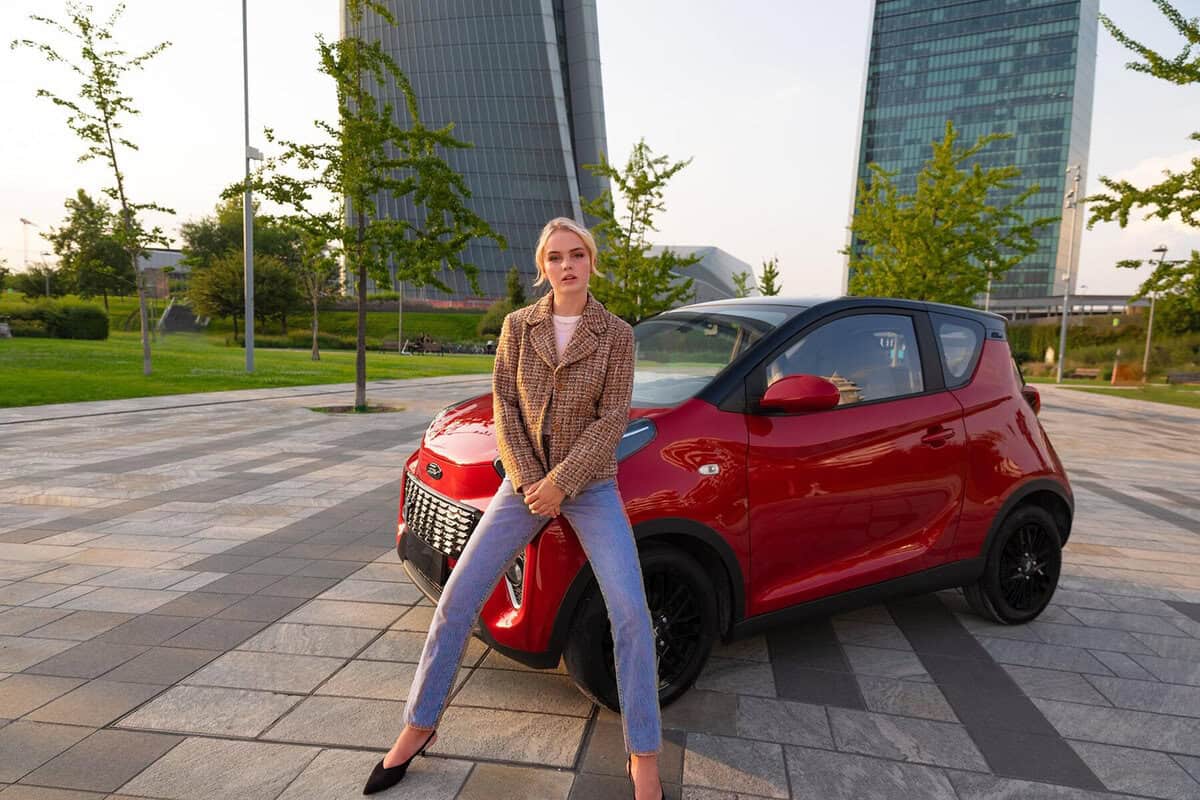 Donna seduta su DR 1.0 EV in piazza con grattacieli sullo sfondo, prospettiva urbana e moderna.