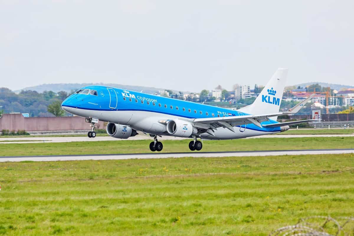 KLM Cityhopper Embraer E-Jet decolla da pista verde, lato destro, ambiente urbano sullo sfondo.