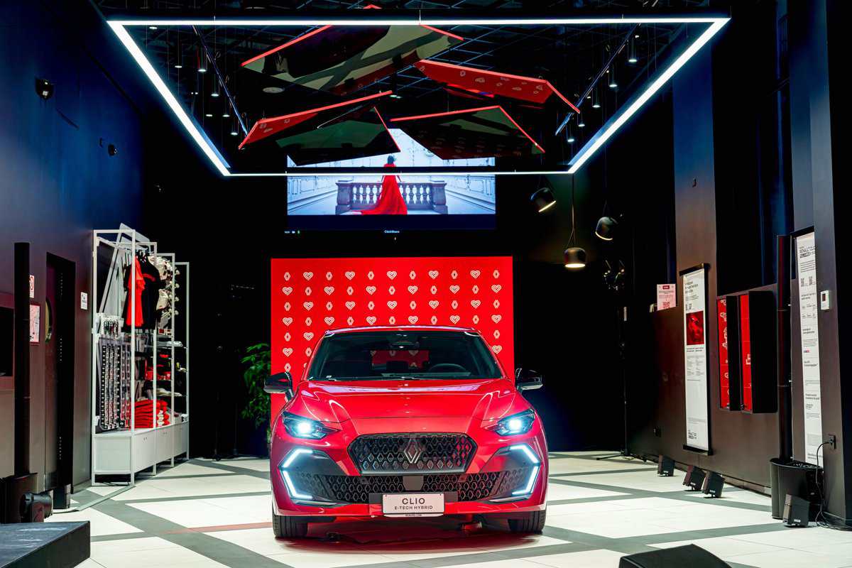 Renault Clio E-Tech Hybrid rossa frontale in showroom, luci accese, sfondo rosso con cuori.