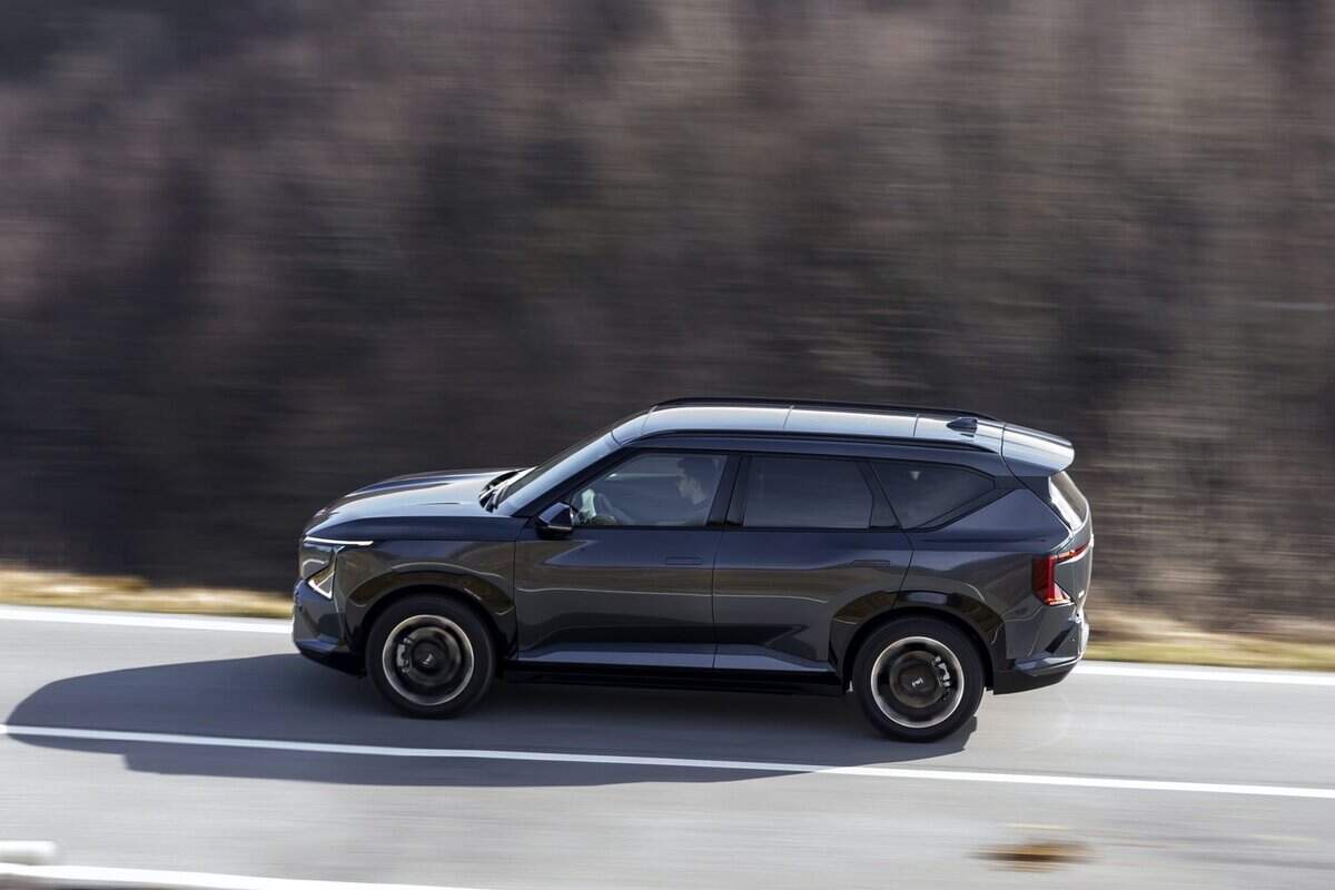 KIA EV5 nera vista laterale su strada, in movimento, su sfondo boschivo.