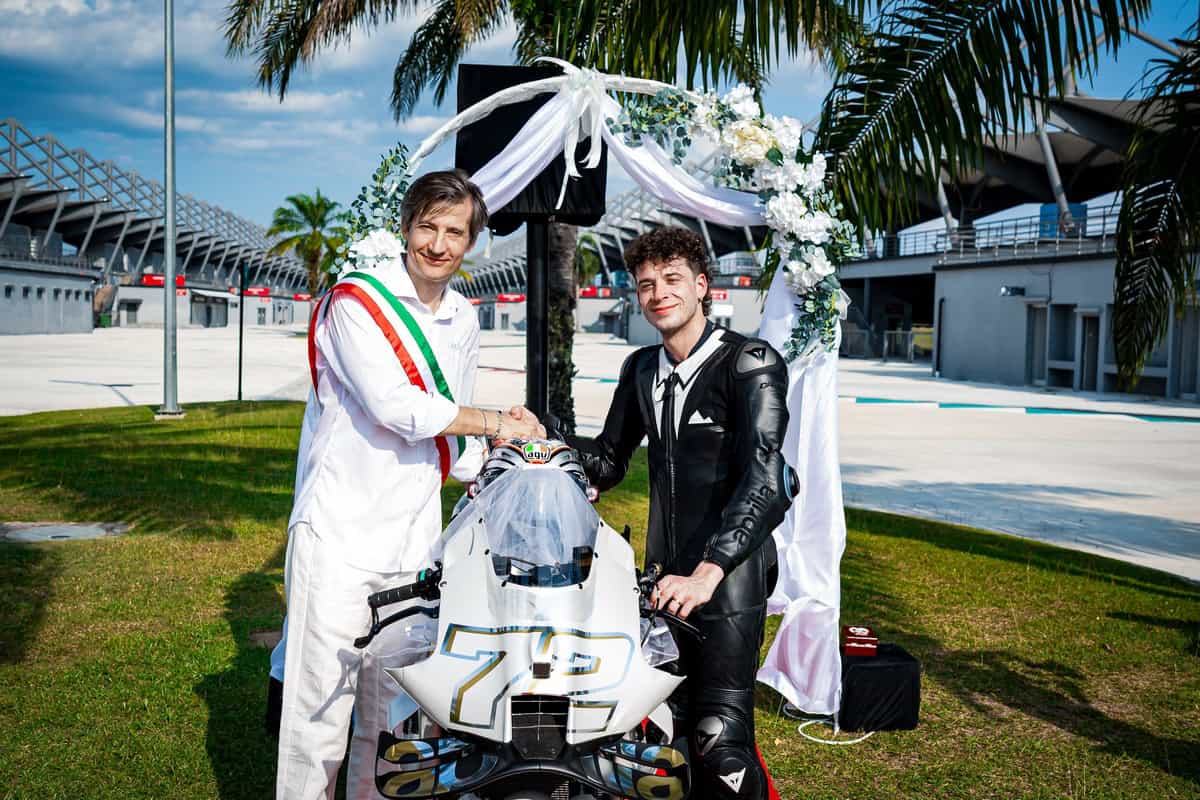 Marco Bezzecchi in tuta nera accanto a ufficiale con fascia tricolore, vicino a una moto bianca, in ambiente esterno.