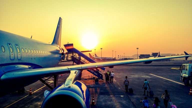 Aereo in partenza all'aeroporto al tramonto con passeggeri che caricano bagagli sullo sfondo.