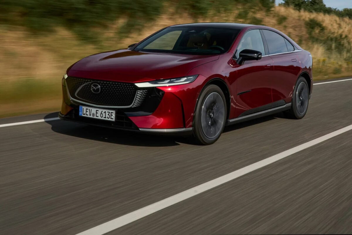 Mazda 6e 2025 rossa in movimento su strada con fondo scuro e vegetazione sullo sfondo.