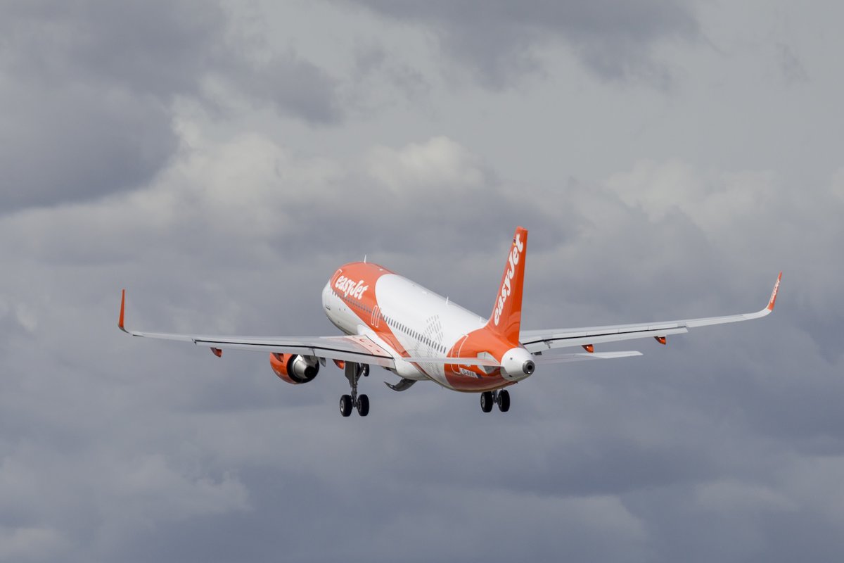 Airbus A320 easyJet arancione e bianco in fase di decollo, ripreso da dietro mentre si alza tra le nuvole grigie.
