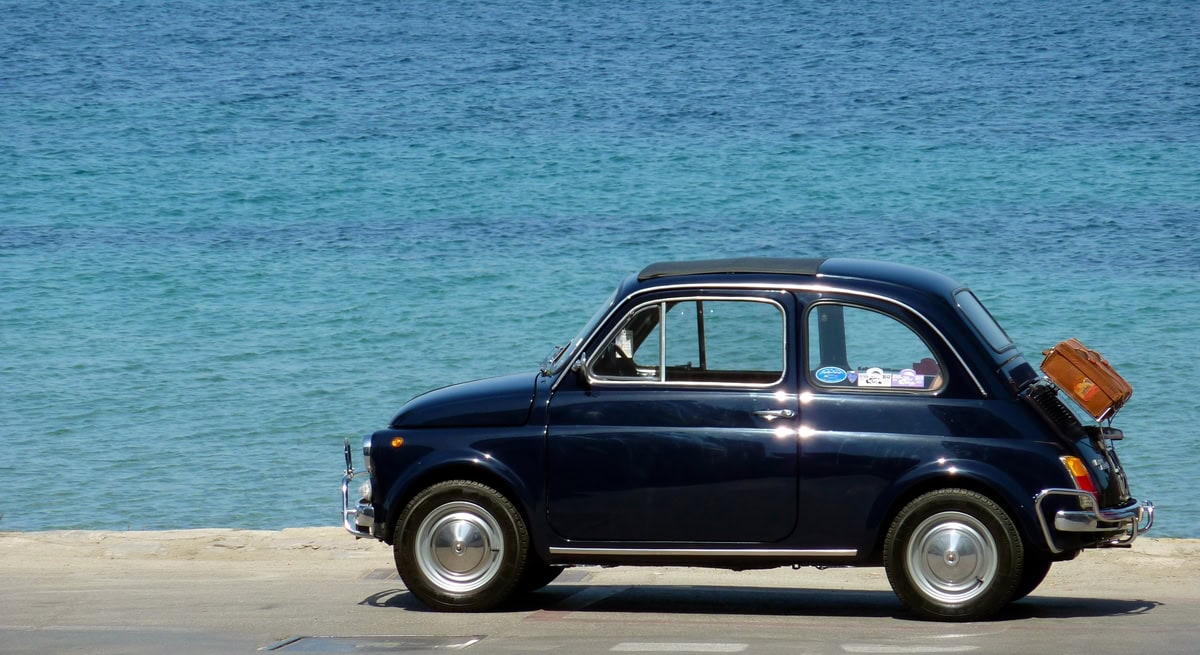Fiat 500 d’epoca blu parcheggiata sul lungomare con valigia sul portapacchi posteriore