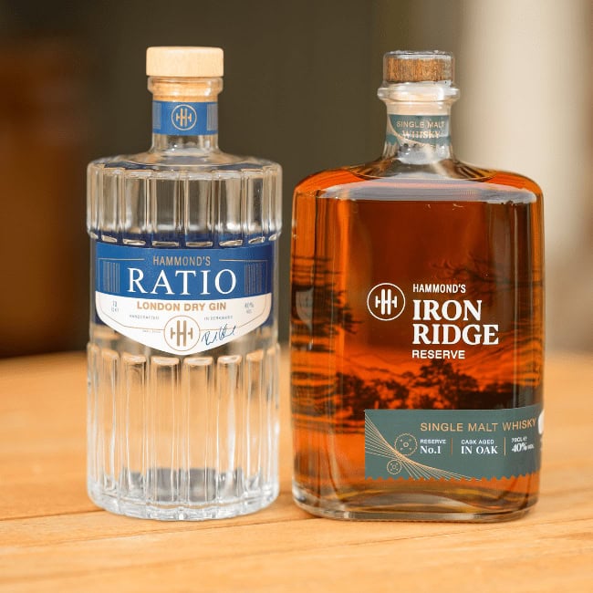 Due bottiglie su un tavolo in legno: a sinistra Hammond’s Ratio London Dry Gin in bottiglia trasparente con etichetta blu, a destra Hammond’s Iron Ridge Reserve Single Malt Whisky in bottiglia ambrata con etichetta scura.