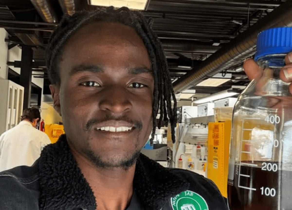 Julian Brown sorride in un laboratorio scientifico mentre mostra un contenitore graduato contenente un liquido scuro, circondato da strumenti da ricerca e attrezzature chimiche.