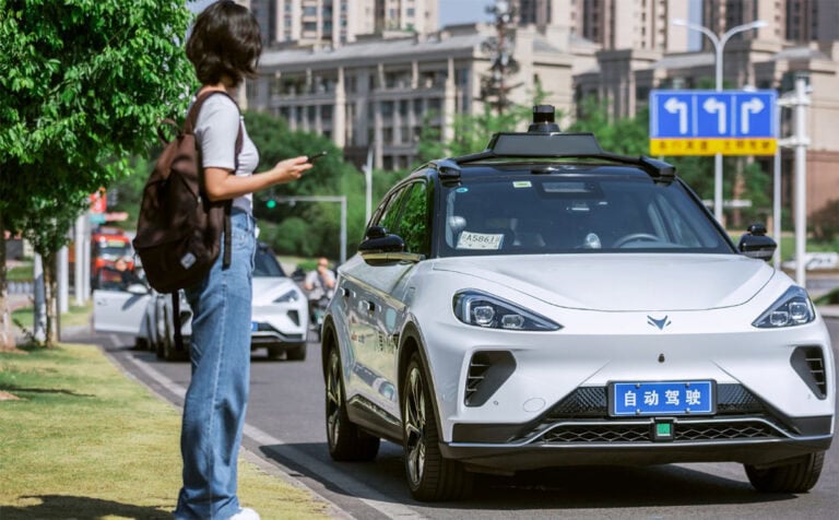 Una giovane donna con zaino si avvicina a un’auto a guida autonoma Pony.ai Apollo Go ferma sul marciapiede, con sensori Lidar sul tetto e targa blu cinese