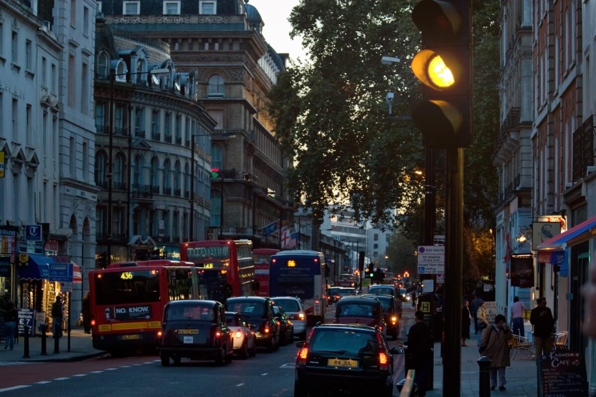 Traffico intenso al tramonto su una strada urbana UK con taxi neri, autobus rossi e semaforo giallo acceso, tipico scenario di Londra con edifici storici e negozi illuminati.