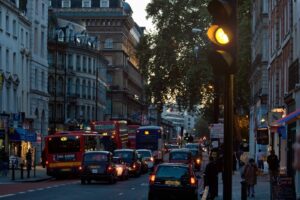 Traffico intenso al tramonto su una strada urbana UK con taxi neri, autobus rossi e semaforo giallo acceso, tipico scenario di Londra con edifici storici e negozi illuminati.