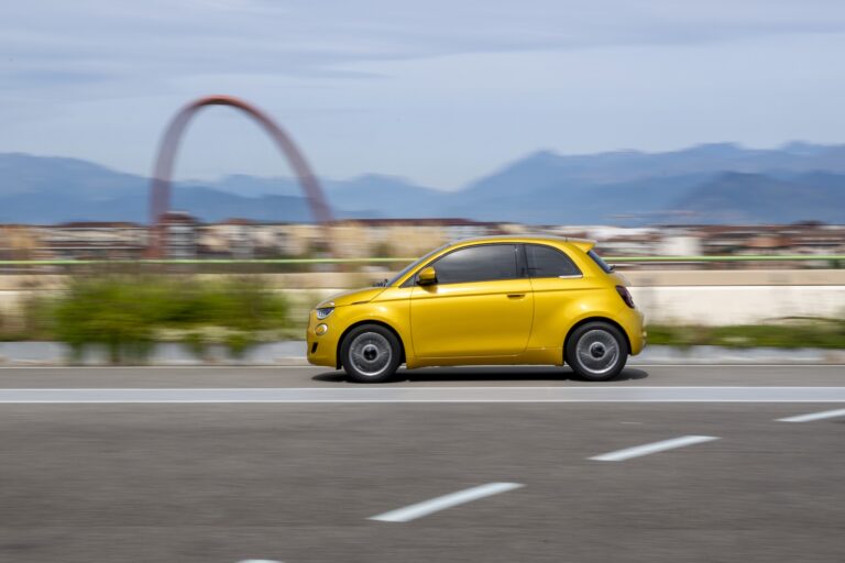 Una Fiat 500 Hybrid gialla in movimento su una strada urbana a Torino, con l’arco olimpico rosso sfocato sullo sfondo.
