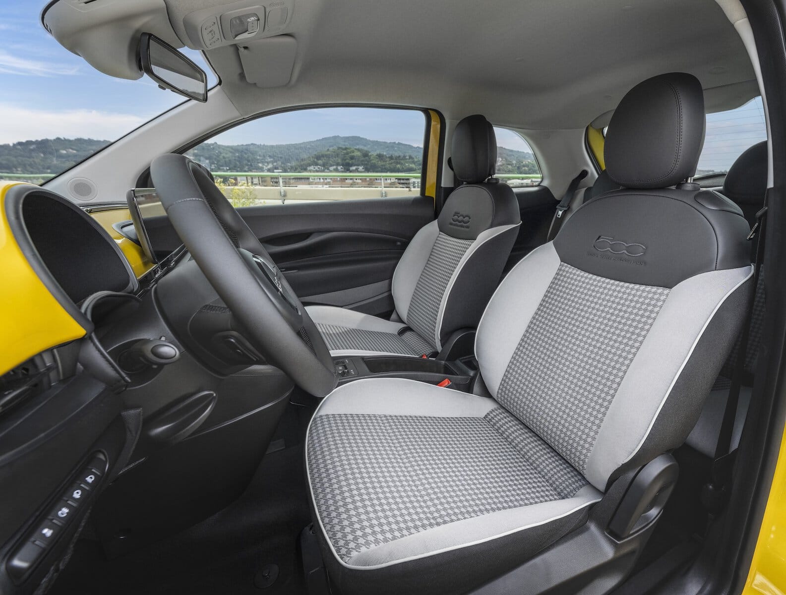 Vista degli interni anteriori di una Fiat 500 Hybrid gialla, con sedili bicolore grigi e neri decorati con il logo 500, parcheggiata con vista collinare.