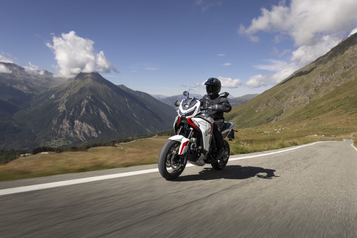 Un motociclista guida la Moto Morini X-Cape 1200 su una strada panoramica in alta quota, con montagne e cielo limpido sullo sfondo.