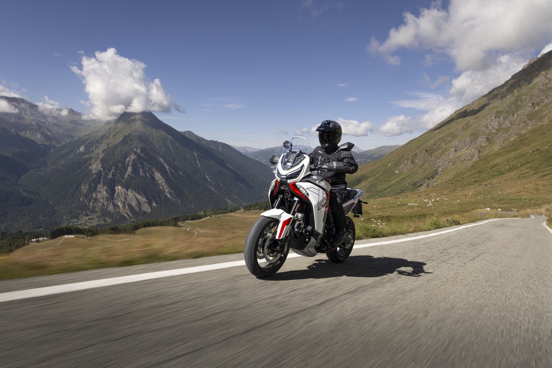 Un motociclista guida la Moto Morini X-Cape 1200 su una strada panoramica in alta quota, con montagne e cielo limpido sullo sfondo.