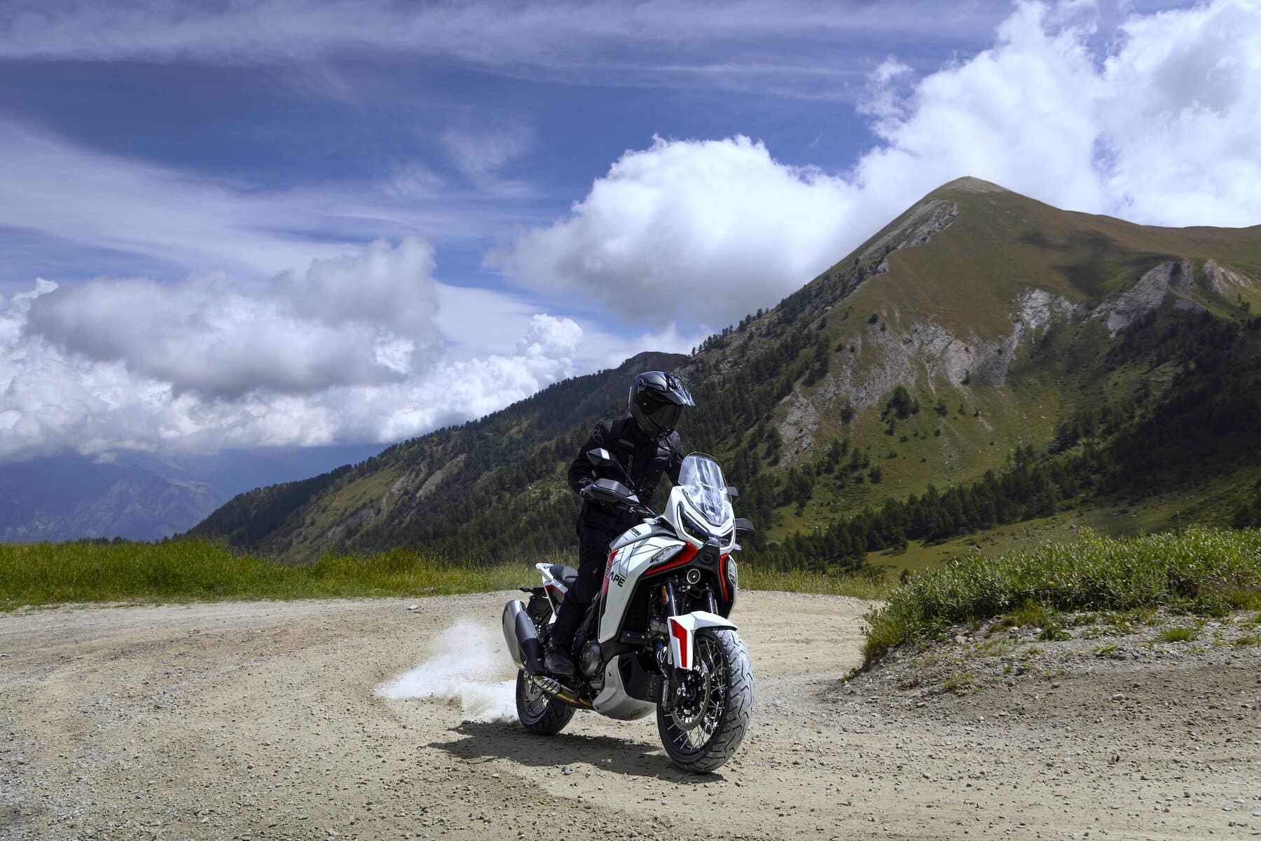 La Moto Morini X-Cape 1200 solleva polvere mentre affronta un tratto sterrato di montagna, circondata da natura e cieli nuvolosi.