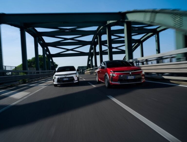 Due modelli di Lancia Ypsilon ibrida, una bianca e una rossa, corrono affiancate su un ponte moderno in acciaio durante una prova su strada. L’immagine mette in risalto il design sportivo e lo stile dinamico della nuova city car ibrida.