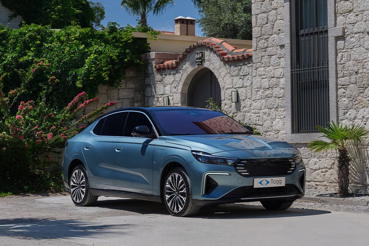 Berlina elettrica Togg T10F color azzurro metallizzato parcheggiata accanto a una villa in pietra con dettagli mediterranei, circondata da piante e fiori sotto il sole estivo.