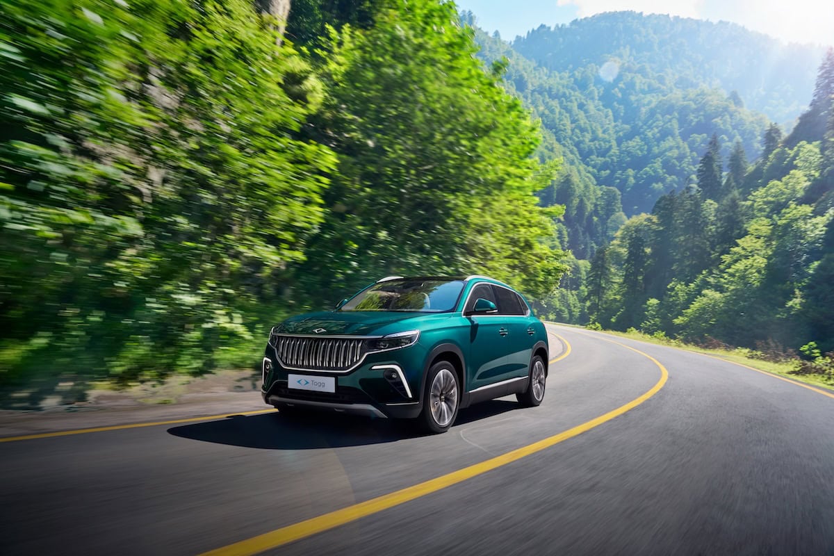 SUV elettrico Togg T10X verde in movimento su una strada di montagna curvilinea circondata da fitti alberi e paesaggio verdeggiante, sotto il sole estivo.