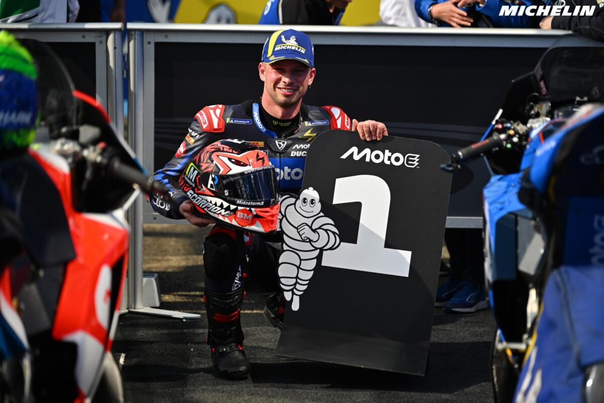 Alt: Pilota di MotoGP sorride e mostra la prima posizione sul podio, indossando tuta da corsa e casco con grafica originale, circondato da moto da gara e staff tecnico.