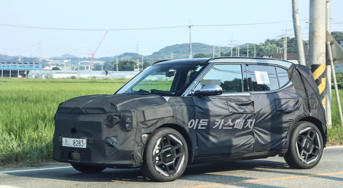 Kia EV2 elettrico in fase di testing, rivestito con camuffamento protettivo, scenario di sviluppo di veicoli green, in viaggio su strada in campagna, anticipando il debutto nel mercato auto elettriche.