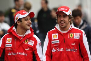 I piloti Ferrari Felipe Massa e Fernando Alonso sorridenti nei box durante un weekend di gara, entrambi in divisa rossa ufficiale con cappellino e loghi degli sponsor.