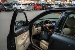 Interni di un'auto parcheggiata con portiera del conducente aperta, vista serale in un parcheggio di supermercato; truffa di una persona che ruba una borsa nera.