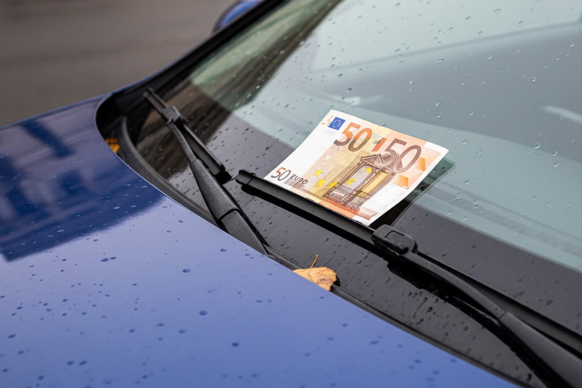Una banconota da 50 euro è bloccata sotto il tergicristallo di un’auto blu parcheggiata sotto la pioggia, con gocce visibili sul parabrezza e sul cofano.